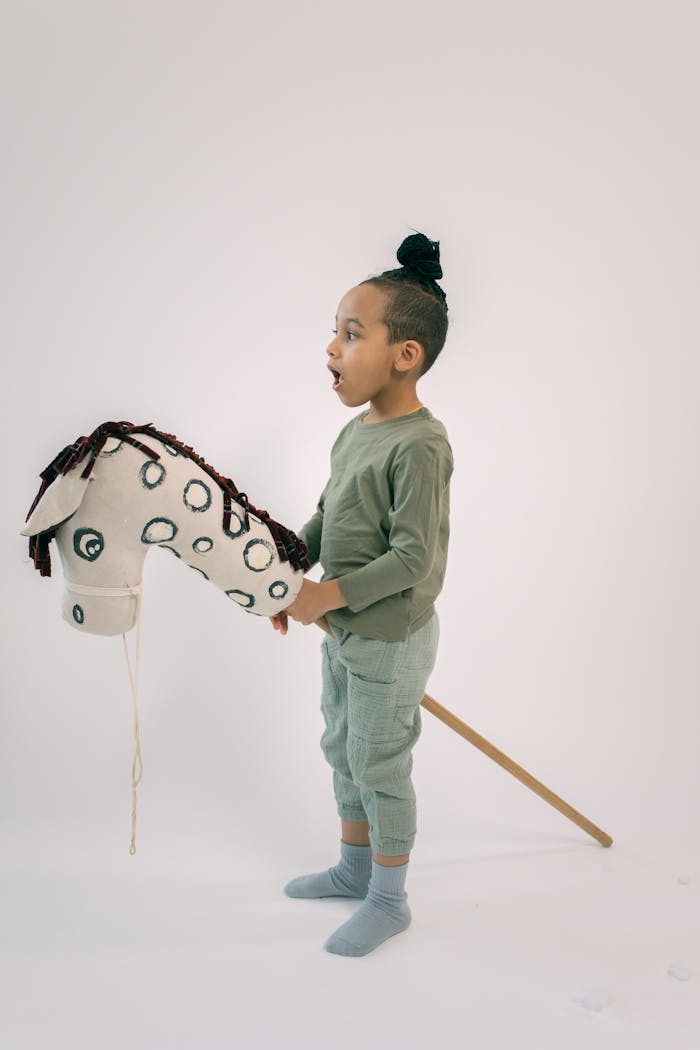 Side view of cute boy in stylish outfit with high bun riding stick horse on white background